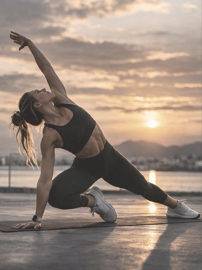 A woman performing a deep side lunge stretch outdoors at sunset in Dubai, training mobility and everyday movement with SHUYO